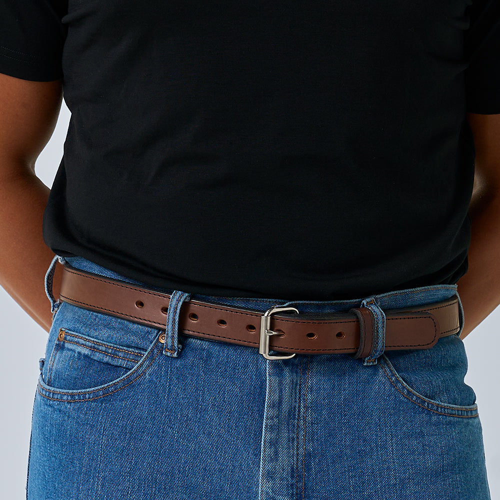 A person stands with their hands behind their back, wearing a black t-shirt, blue jeans, and The Xtreme - 1.25&quot; brown concealed carry belt by Hanks Belts with a silver buckle.