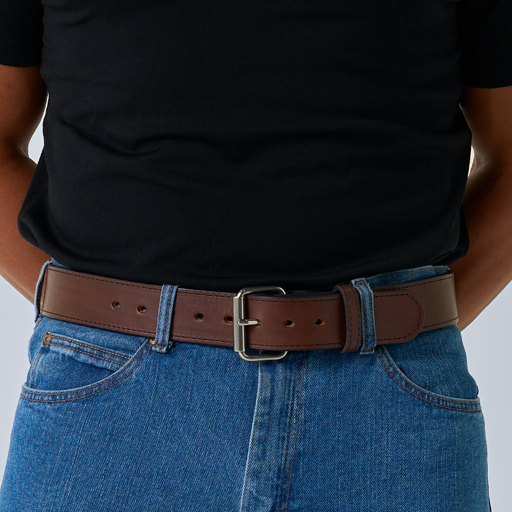 Dressed in a black t-shirt tucked into blue jeans, they wear The Xtreme - 1.75&quot; heavy duty leather work belt from Hanks Belts, featuring a silver buckle, with their hands behind their back.