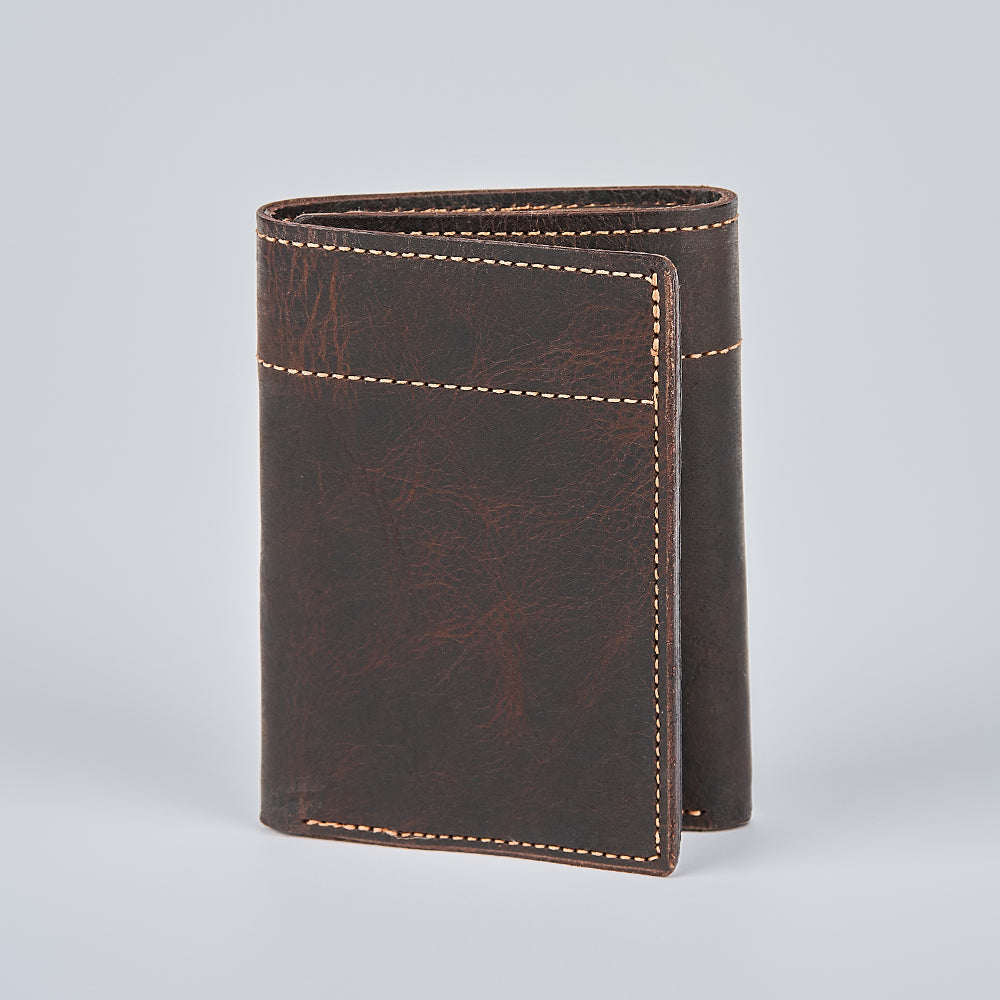 The Hanks Belts Fontana Bison Trifold, a handcrafted dark brown bison leather wallet with tan stitching, stands upright on a plain light gray background.