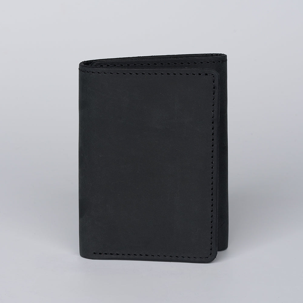 The Saranac Premium Trifold by Hanks Belts, a black minimalist American-made wallet, stands upright on a plain light gray background, closed to display its neat edge stitching.