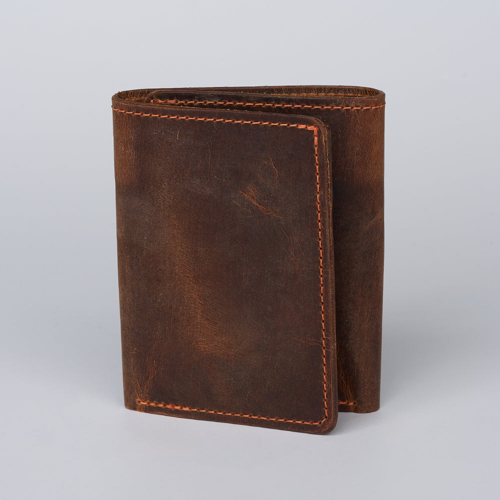A Hanks Belts Saranac Premium Trifold wallet with visible stitching stands upright on a light gray background. Made in the USA, this wallet has a rugged, rustic look and is shown partially folded closed.
