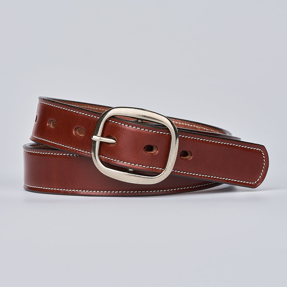 The Eloise by Hanks Belts—a women’s vegetable-tanned leather belt with white stitching and a silver oval buckle—is coiled on a light gray background.