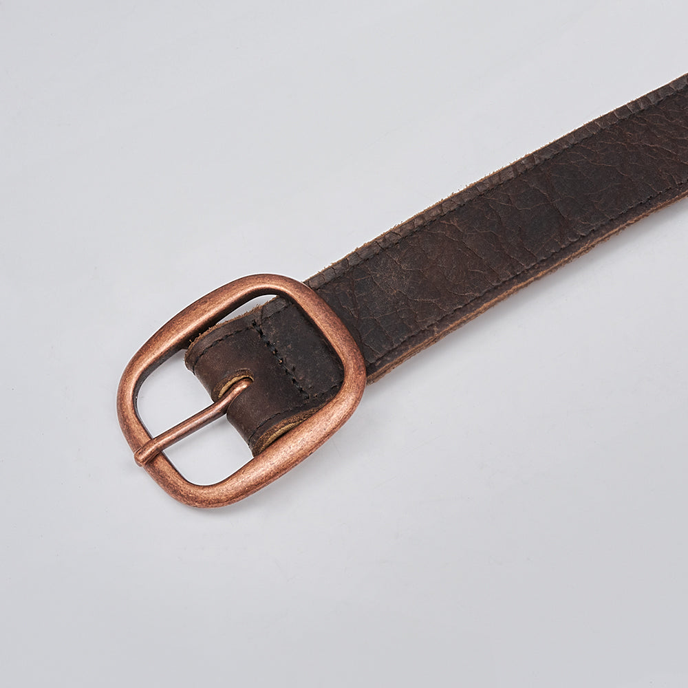 A close-up of the Ember - 1.25&quot; brown leather belt by Hanks Belts, featuring a worn, textured surface and a copper-colored metal buckle against a light gray background.