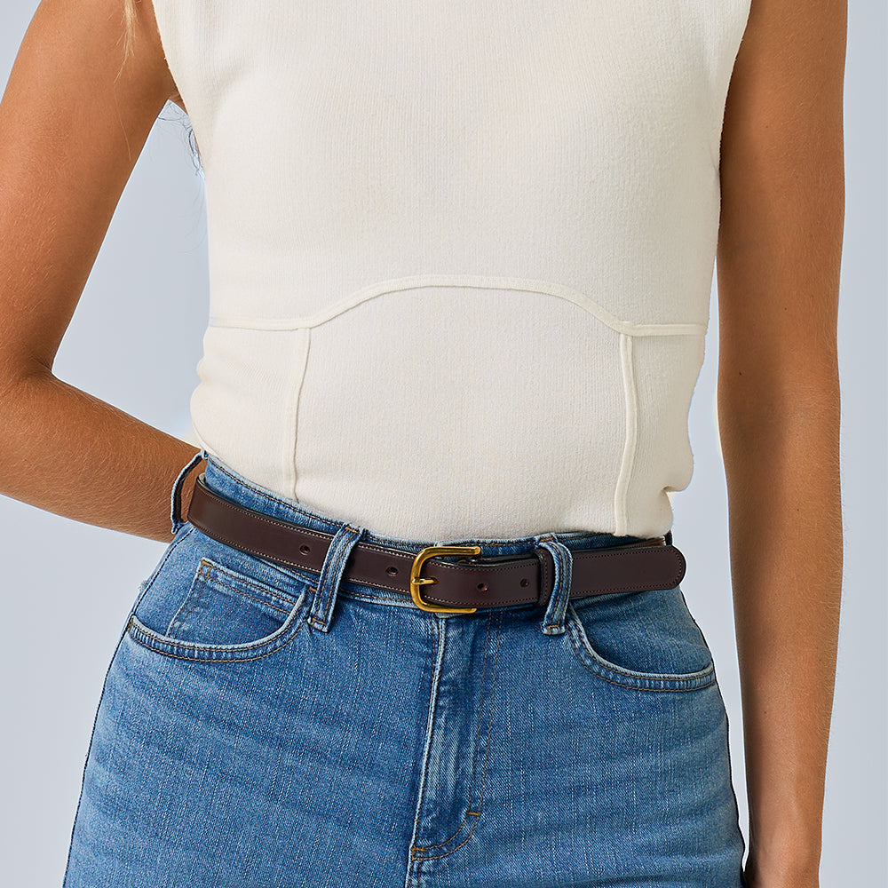 A person wears a sleeveless cream top with high-waisted blue jeans and the Addyson - 1&quot; leather belt by Hanks Belts, featuring a brass buckle. The photo is cropped at the shoulders and mid-thigh.