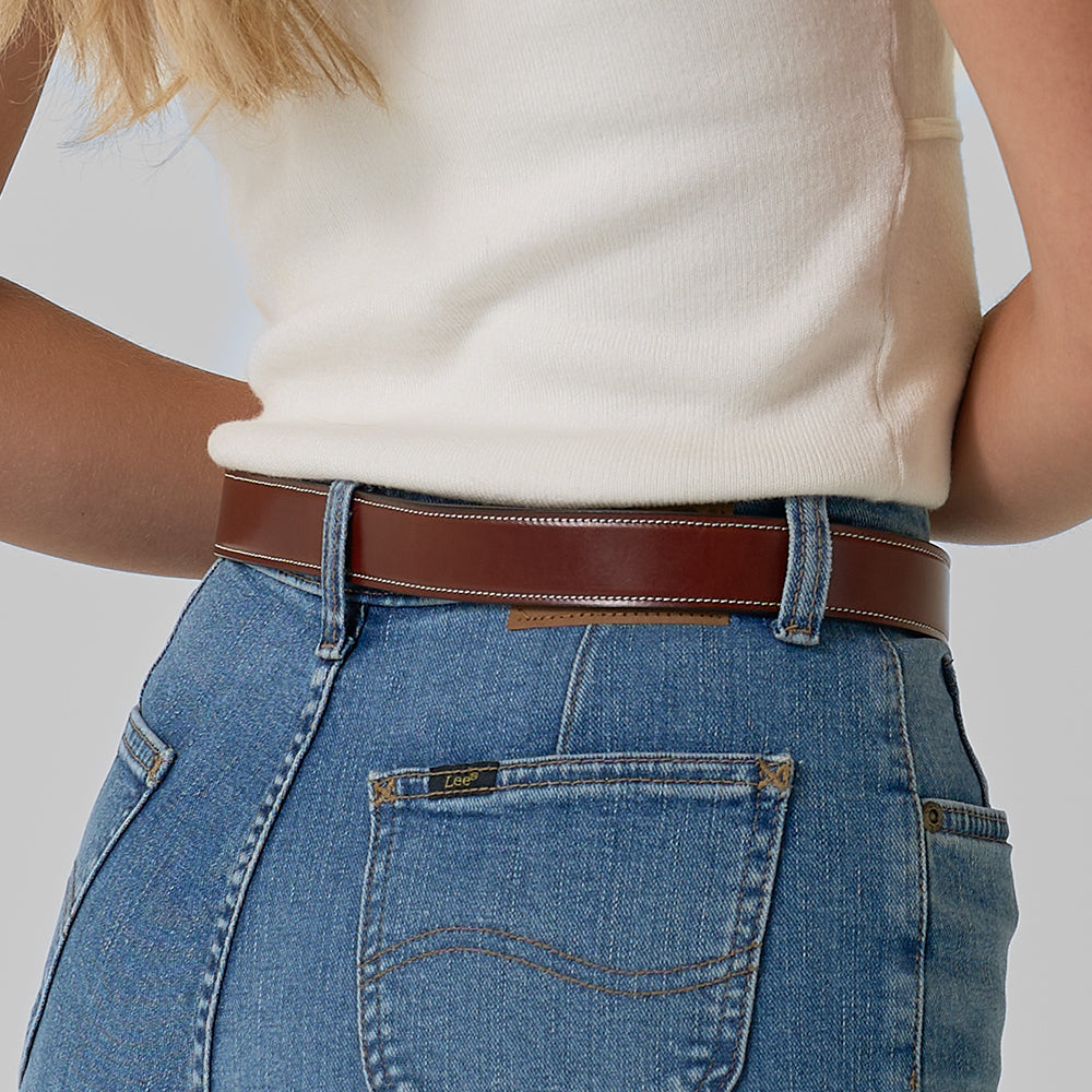 A person shown from the waist down and slightly from the side wears a sleeveless white top with high-waisted blue jeans, styled with The Eloise—Hanks Belts’ women&#39;s vegetable-tanned leather belt.