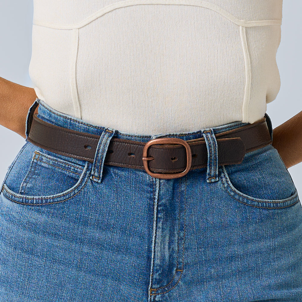 A person in blue high-waisted jeans styles a cream fitted top with Hanks Belts’ Ember - 1.25&quot;, a dark brown leather belt featuring a rounded buckle.