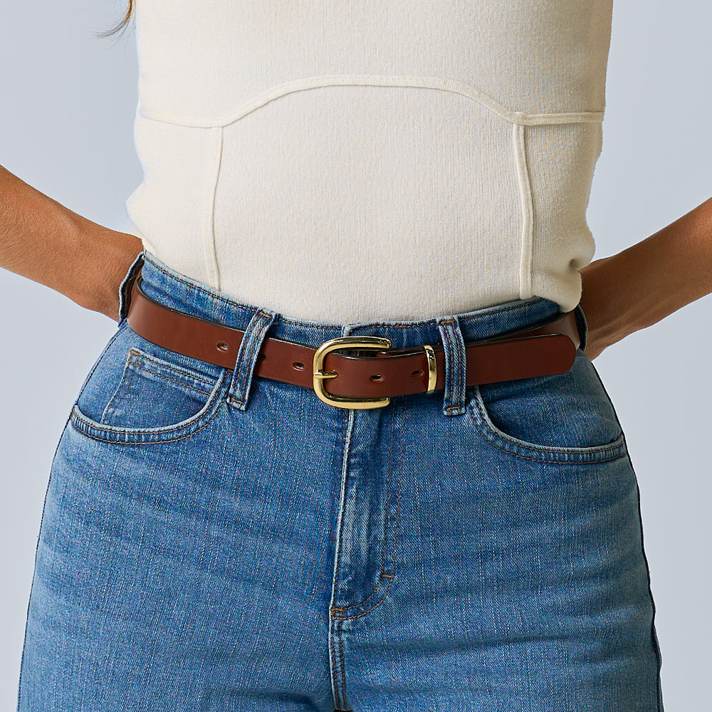 A person wears a cream top tucked into blue jeans with The Poppy women&#39;s statement belt by Hanks Belts, featuring oak leather and a brass buckle. The image is cropped to show only the torso and waist.