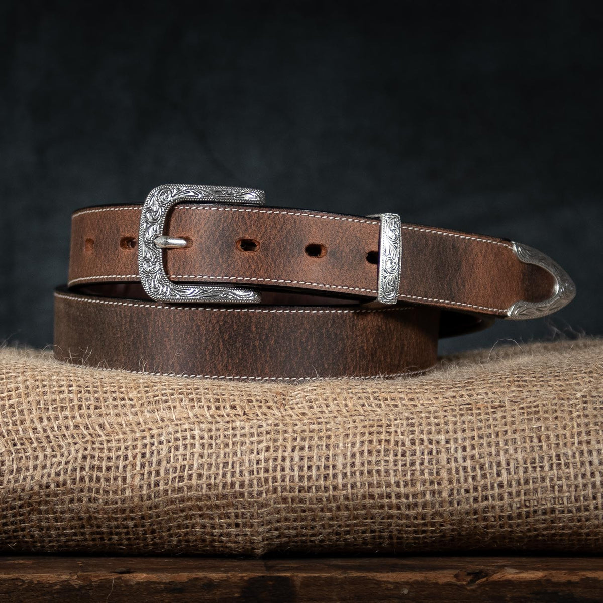 A brown leather Willow Belt - 1.25&quot; by Hanks Belts, designed for women with a silver floral buckle and tip adorned with engraved details, is coiled and placed on a piece of burlap fabric against a dark background. This adjustable belt, featuring multiple holes for a perfect fit, comes with an impressive 100-year warranty.