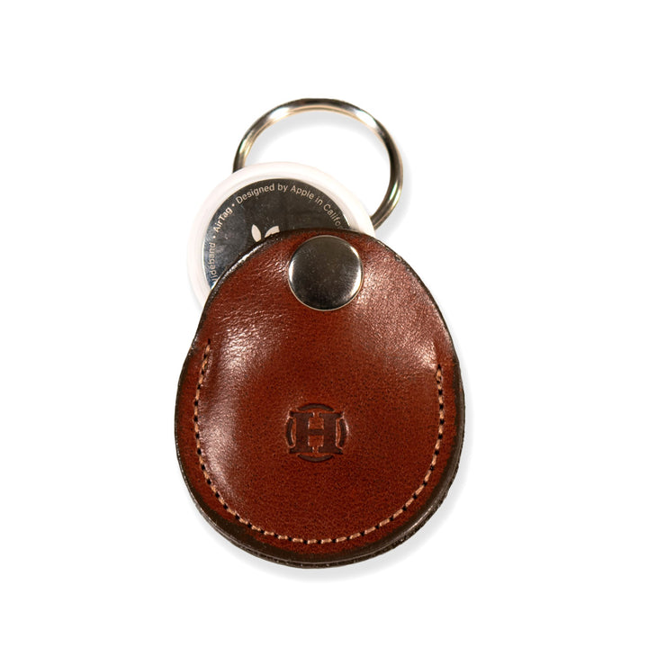 Introducing the Circular Tag Holder by Hanks Belts: This oak leather keychain pouch, crafted from USA-tanned full-grain hide, features a secure snap closure adorned with a silver button and a sturdy metal keyring. Embossed with an "H," it serves as an elegant AirTag leather holder, showcasing the round white device above the pouch.