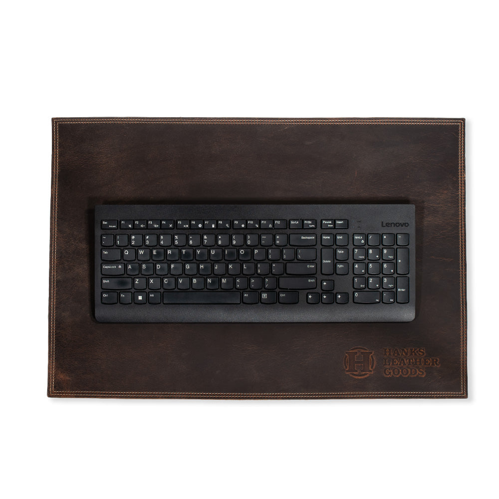 A black Lenovo keyboard sits atop a dark brown desk pad crafted from premium bison leather. This handmade piece, produced in the USA, showcases visible stitching and is embossed with "Hanks Belts" along with a decorative emblem in the bottom right corner.