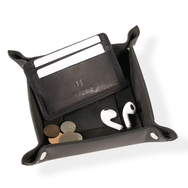 A black Italian Large Valet Tray from Hanks Belts, crafted from full-grain leather and embossed with "Hawes," stylishly holds a sleek cardholder with three white cards, coins, and white wireless earbuds. It features reinforced corners for travel-ready convenience.