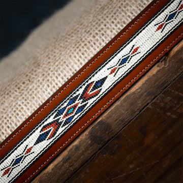 A close-up showcases The Seneca - 1.5" by Hanks Belts, a brown leather belt made from premium English Bridle Leather with diamond-shaped, multicolored geometric embroidery. It exudes western spirit, resting on textured burlap on a dark wooden surface.