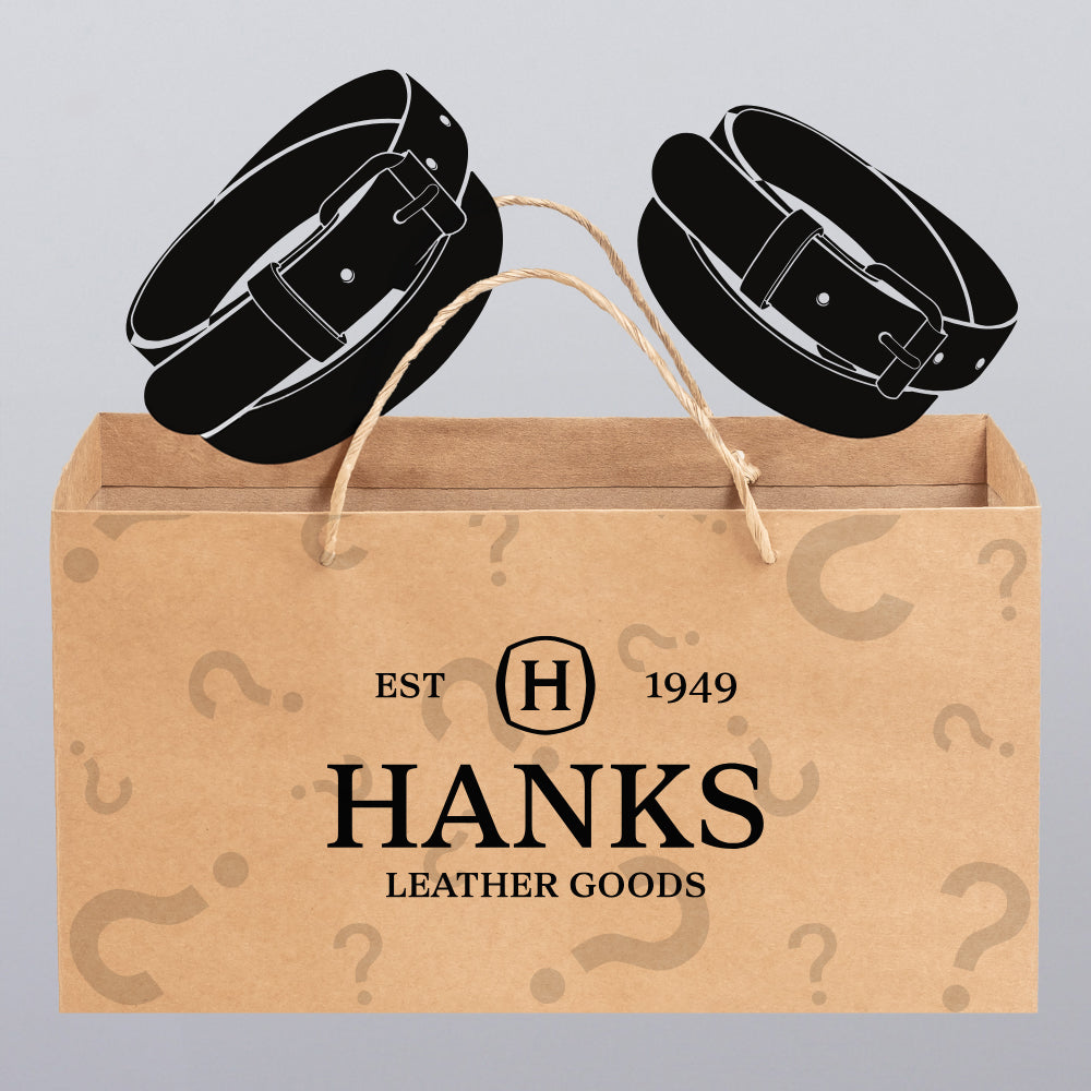 A brown paper bag labeled "Hanks Belts" and "EST 1949" is decorated with question marks, with two black belts showing. Inside is the Less-Than-Perfect Mystery Bag: 2 Belts Per Bag from Hanks Belts.