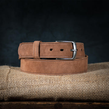 The Suede Belt - 1.5" by Hanks Belts, made in the USA from fine nubuck leather, is neatly coiled on burlap. It features a silver buckle and adjustable sizing with multiple holes. A dark background highlights the rich textures of both belt and cloth.