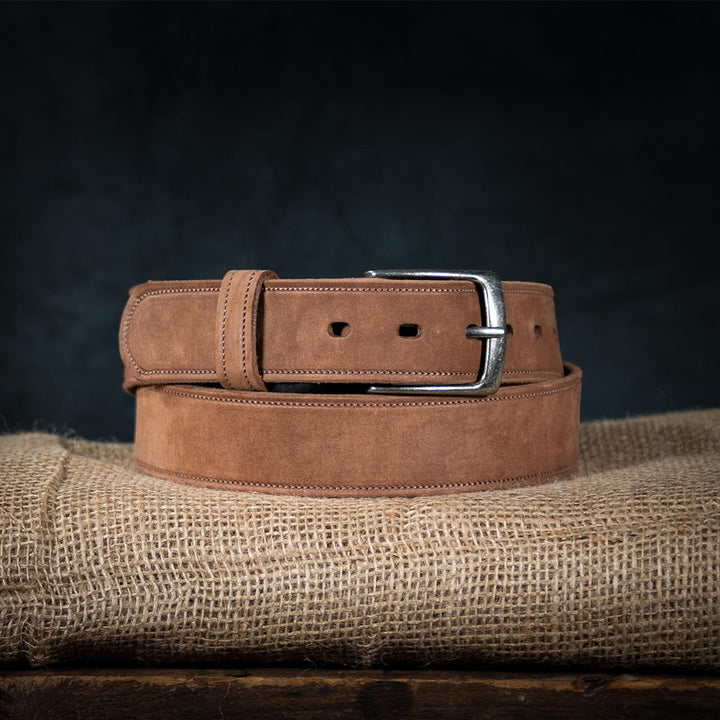 The Suede Belt - 1.5" by Hanks Belts, made in the USA from fine nubuck leather, is neatly coiled on burlap. It features a silver buckle and adjustable sizing with multiple holes. A dark background highlights the rich textures of both belt and cloth.