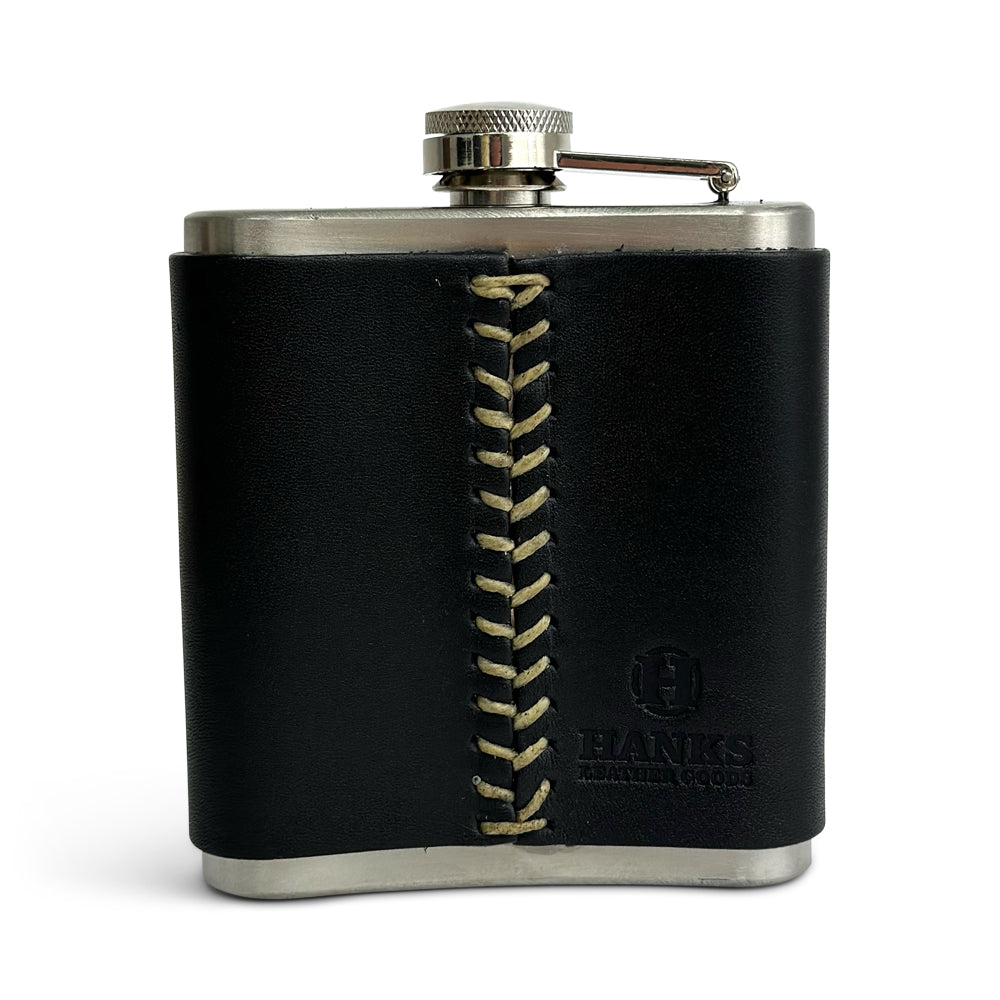 The Leather Covered Flask by Hanks Belts features a stainless steel body wrapped in USA tanned leather, highlighted by a unique vertical stitch pattern in the front center. The leather wrap also showcases the &quot;HANKS&quot; logo and text embossed at the bottom right corner. This flask comes equipped with a screw-on cap secured by a metal hinge.