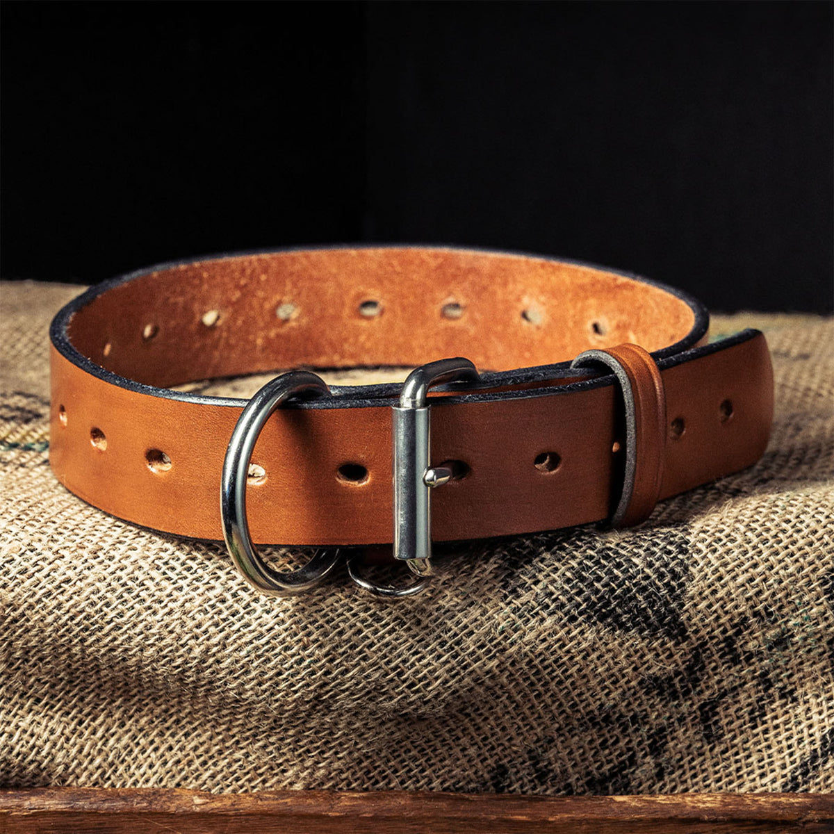 The Big Ass Dog Collars for Big Ass Dogs by Hanks Belts is a brown leather heavy-duty collar with a metal buckle and D-ring, shown on textured burlap against a dark background.