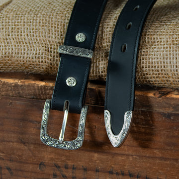 The Dakota Belt by Hanks Belts is made from North American bison leather and features an ornate silver floral buckle and tip. The intricate engraved designs add a decorative touch, displayed on burlap over a wooden surface.
