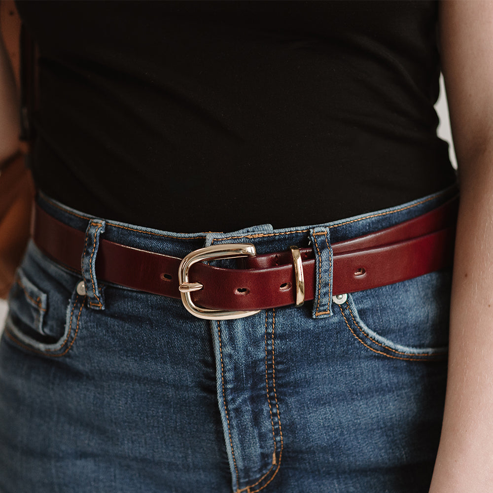 A person wears a black shirt tucked into blue jeans, secured with Hanks Belts&#39; The Poppy - 1&quot;, a premium leather belt with a shiny brown finish and gold buckle, offering versatile style.