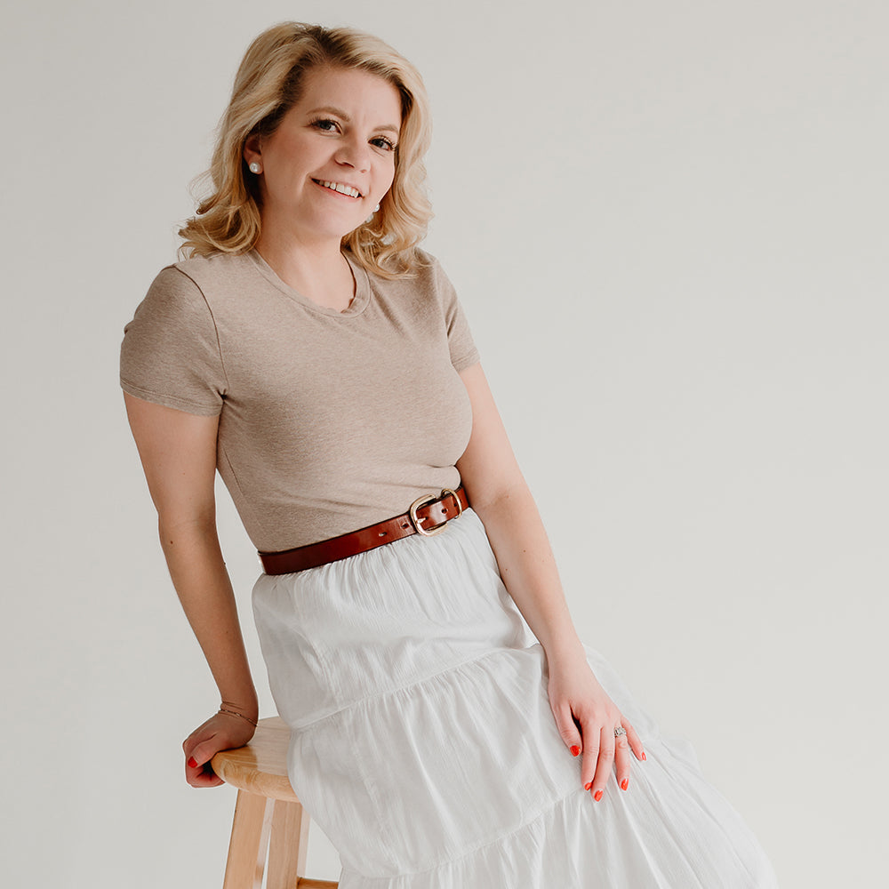 A blonde woman in a beige t-shirt and white skirt sits on a wooden stool against a plain light background, smiling at the camera and wearing The Poppy - 1&quot; by Hanks Belts, a premium leather belt and versatile accessory.
