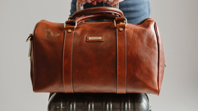 model standing with hanks leather duffel on top of a suitcase rolling it