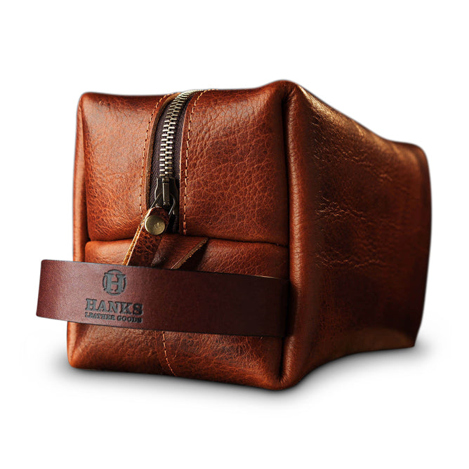 Introducing the Bison Leather Dopp Bag by Hanks Belts: a handcrafted brown leather Dopp kit made in the USA, featuring a brass zipper and a convenient carrying strap. The strap is adorned with "HANKS LEATHER GOODS" text and a logo imprint. Constructed from premium USA Bison Leather, this bag boasts a textured finish, set against a white background.