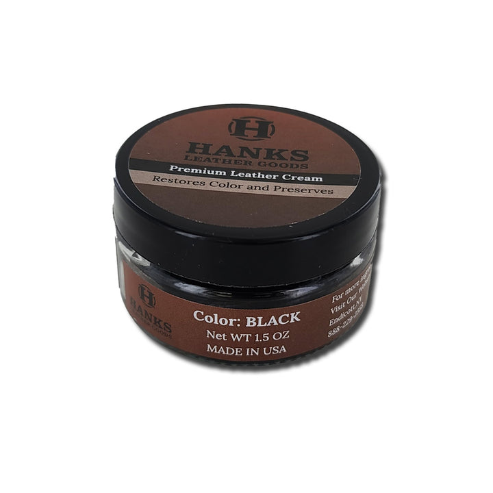 A small round jar of Hanks Belts Premium Leather Cream and Color Restoration. The predominantly brown container with a black lid features a label indicating it restores color and preserves leather. The 1.5 oz, USA-made jar contains black premium leather cream to revive and refinish your leather products.