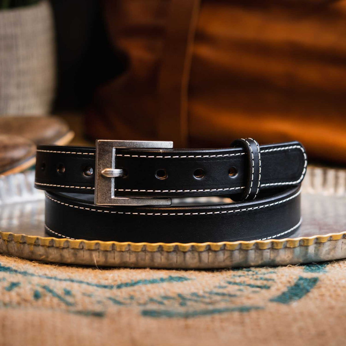 A durable Bonnie Belt - 1.25&quot; from Hanks Belts, with white stitching along the edges, is coiled neatly on a decorative metal tray. This women&#39;s gun belt features a silver rectangular buckle and several holes for adjustment. The background showcases a blurred brown bag and some fabric.
