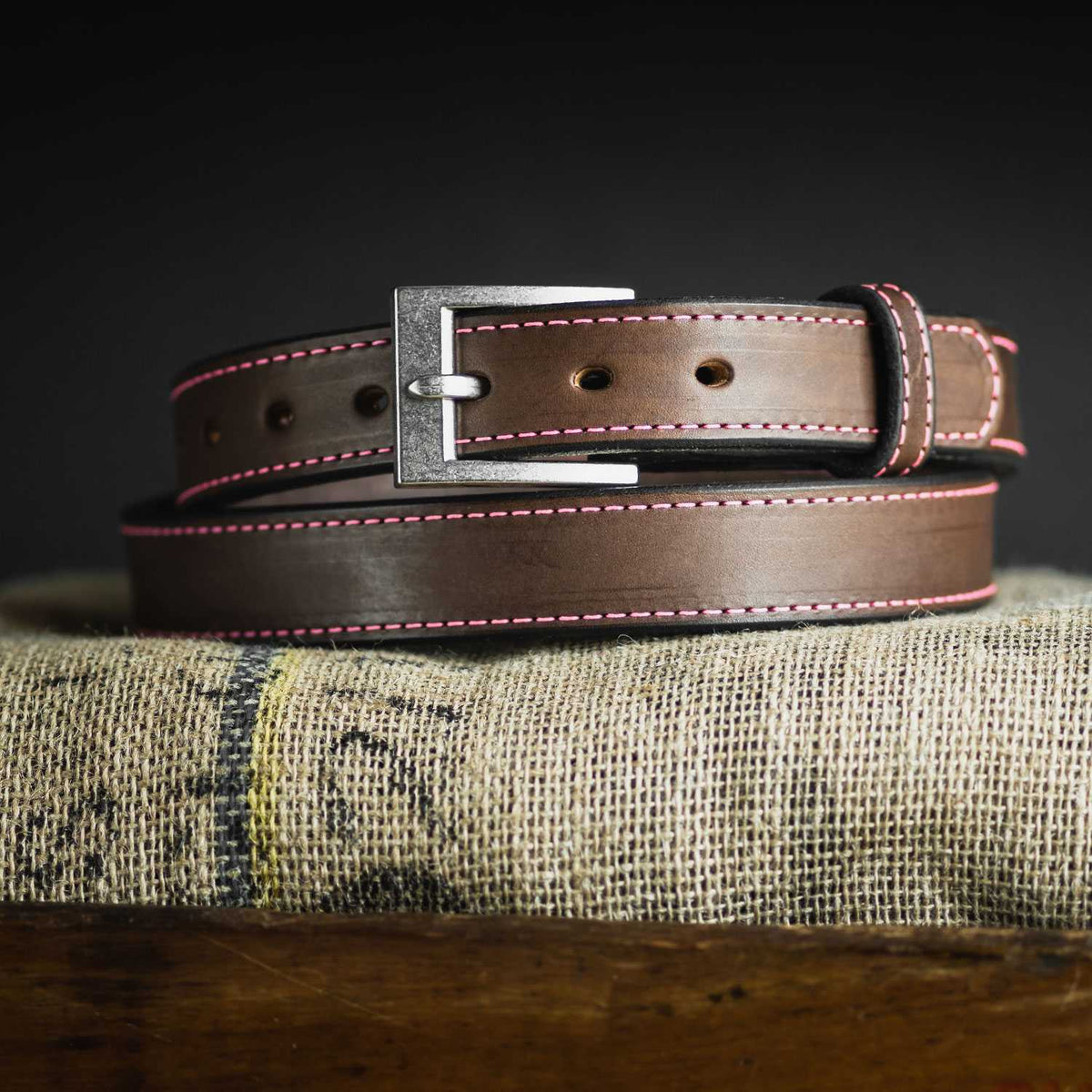The Bonnie Belt - 1.25&quot; by Hanks Belts, a women&#39;s gun belt in brown leather with pink stitching and a silver buckle, is coiled and displayed on a piece of burlap fabric. The rustic setting accentuates the craftsmanship and design details of this CCW belt against a dark background.