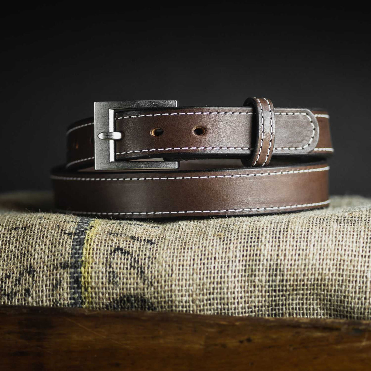 The Bonnie Belt - 1.25&quot; by Hanks Belts, a brown women&#39;s gun belt adorned with white stitching, is coiled and displayed on a burlap sack. This durable leather belt features a metallic buckle and is set against a dark background, highlighting its craftsmanship and texture.