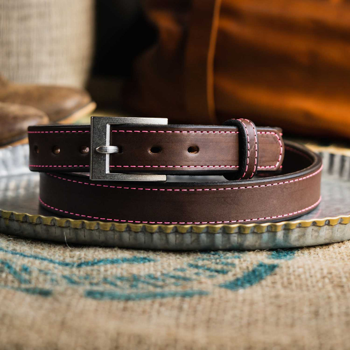The Bonnie Belt - 1.25&quot; by Hanks Belts, in dark brown with pink stitching and a silver buckle, is coiled neatly on a galvanized metal tray. The background reveals a blurred, rustic setting with a bag and partially visible brown boots.