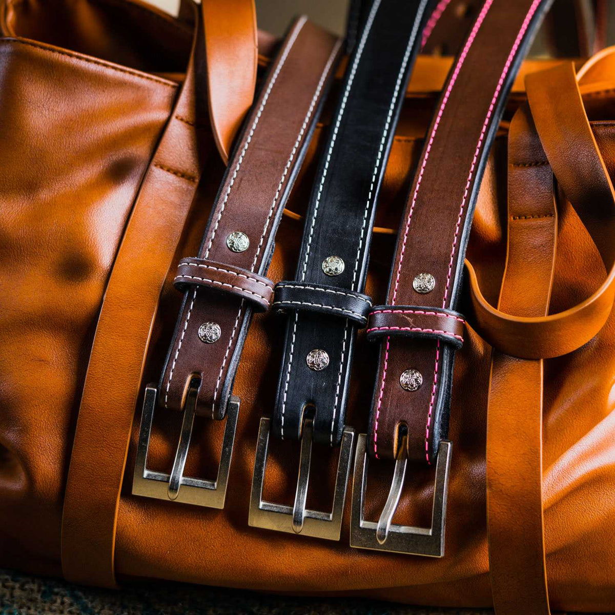 A close-up shot of three Hanks Belts&#39; Bonnie Belt - 1.25&quot; with metal buckles lying on a brown leather bag. The belts are in brown, black, and tan colors, each featuring contrast stitching and shiny, circular metal studs.