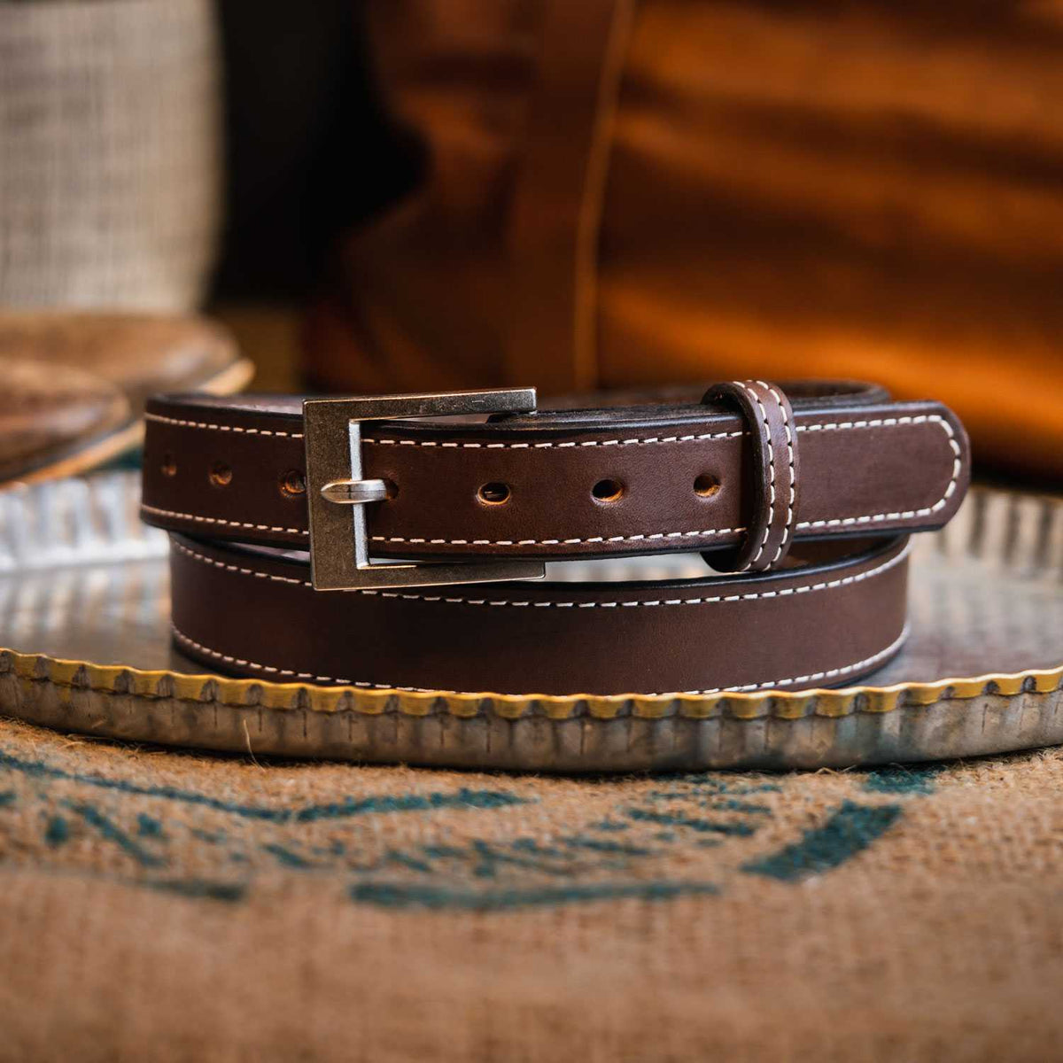 The Bonnie Belt - 1.25&quot; by Hanks Belts, a durable leather belt in brown with white stitching, is coiled and displayed on a metal tray. Featuring a silver buckle and multiple adjustment holes, the belt is presented on a textured fabric surface.