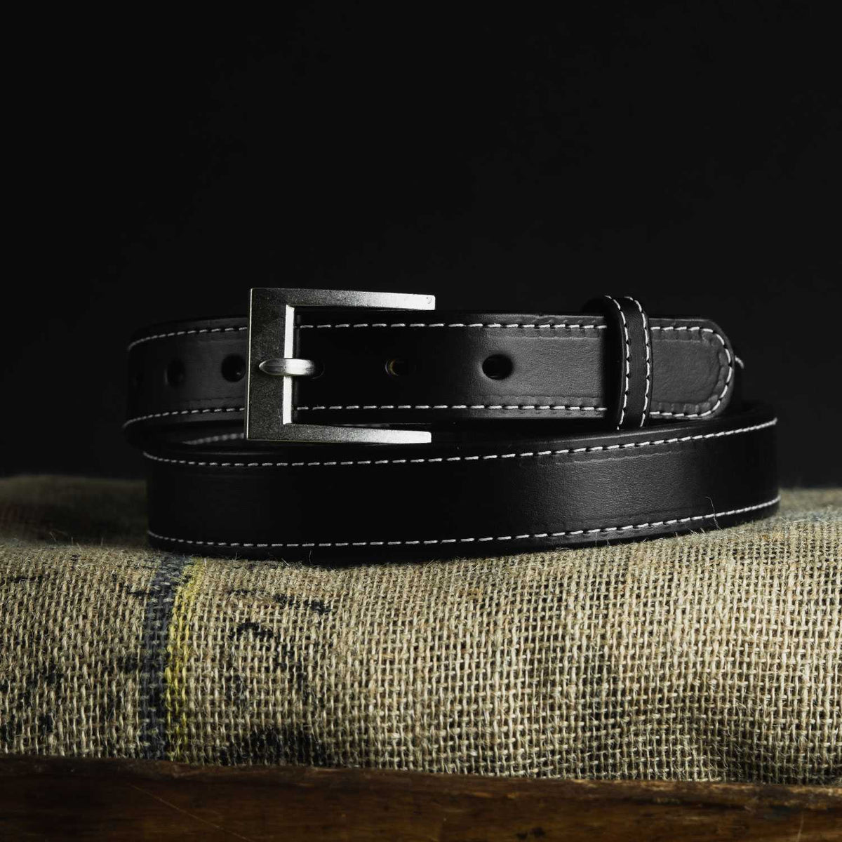 A Bonnie Belt - 1.25&quot; from Hanks Belts, crafted in black leather with white stitching and featuring a silver buckle, coiled neatly and placed on a textured burlap cloth against a black background.