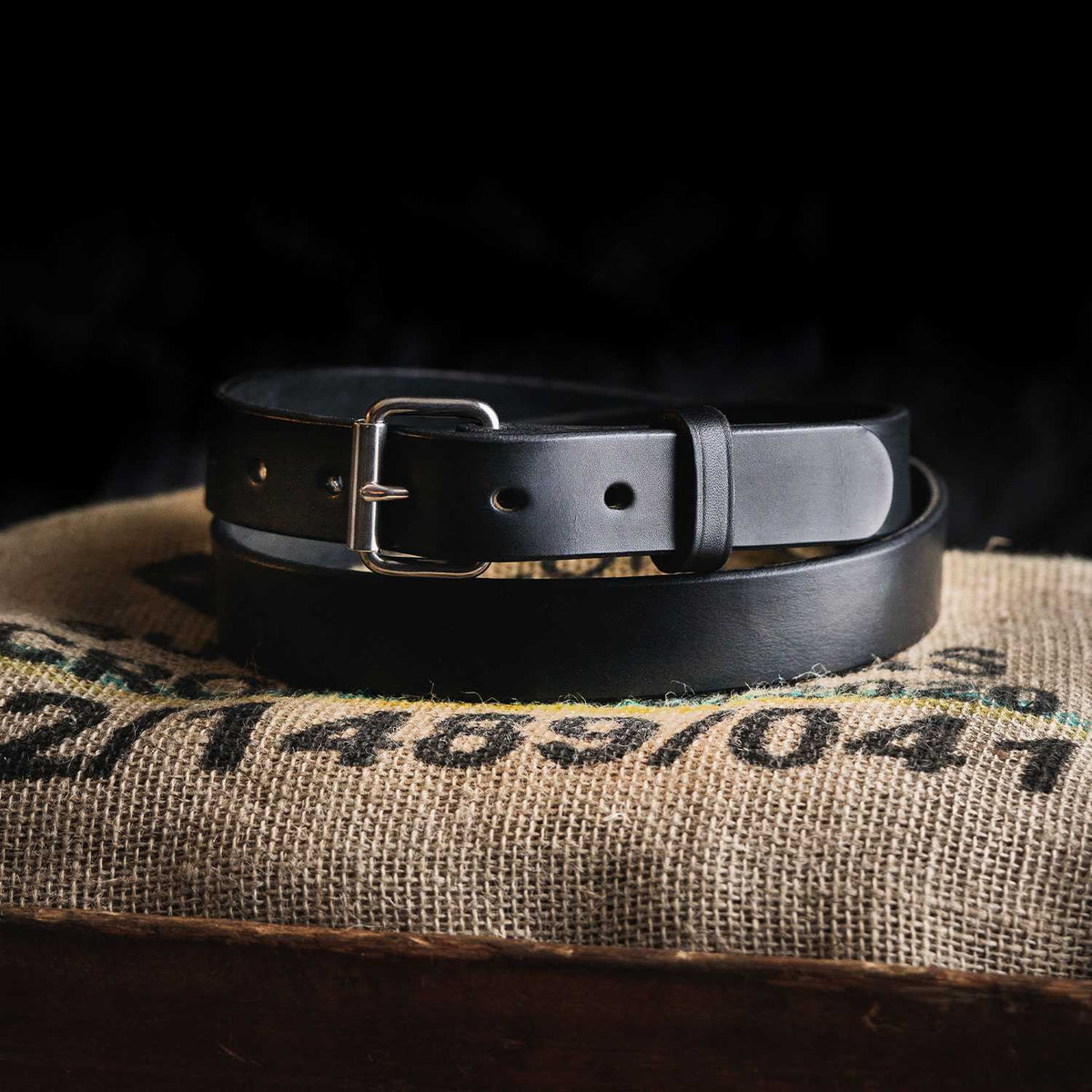 A black, durable leather Deputy Belt from Hanks Belts, featuring a silver buckle, is neatly coiled on top of a burlap sack. The dark background makes both the belt and sack stand out prominently. This 1.25&quot; wide belt showcases a simple, classic design with several adjustment holes.