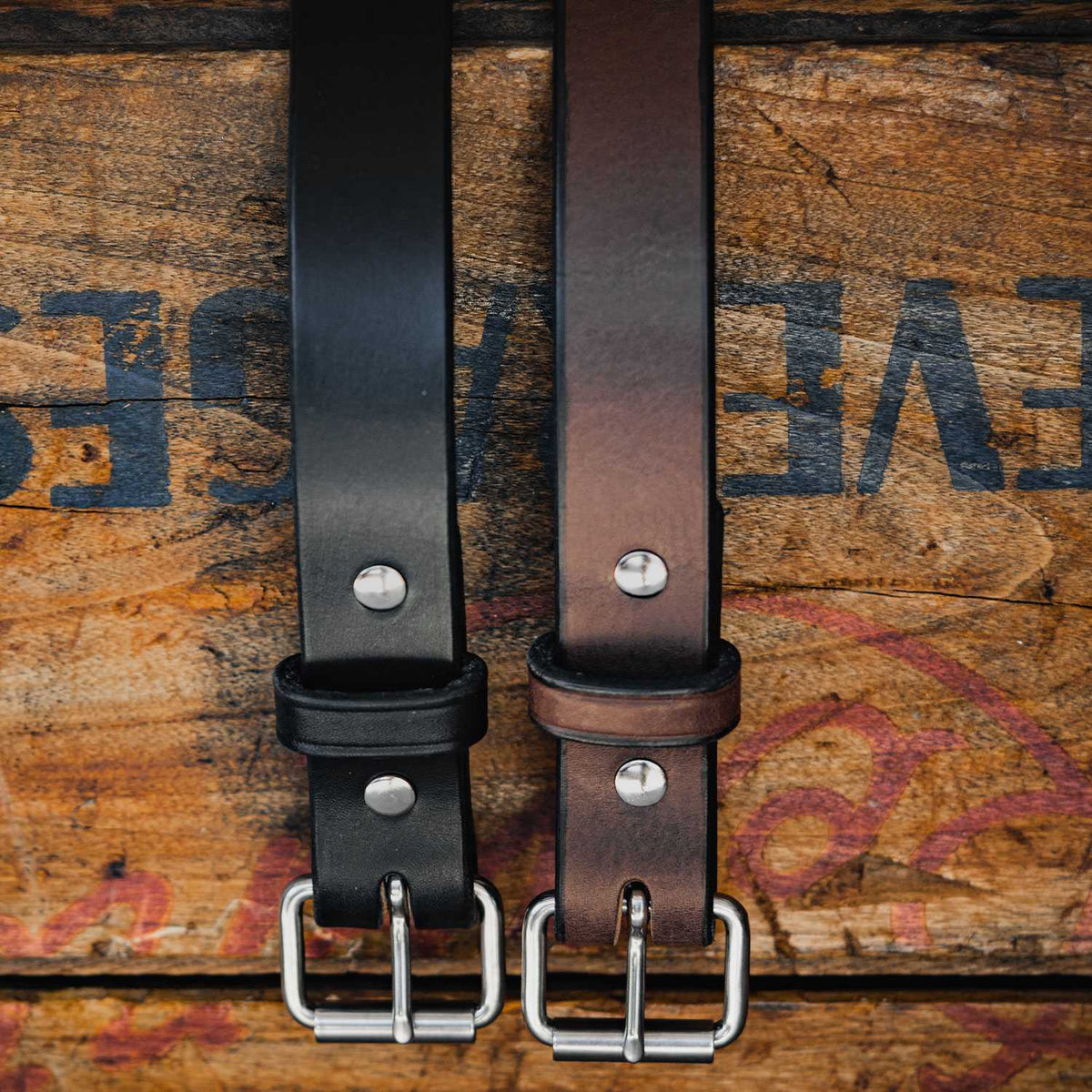 Two durable leather Hanks Belts Deputy belts, one black and one brown, are laid out side by side on a rustic wooden surface. Both belts feature silver buckles and two metal rivets near the buckle. The 1.25&quot; wide Deputy belts complement the wood background with its faint, partially visible black and red markings.