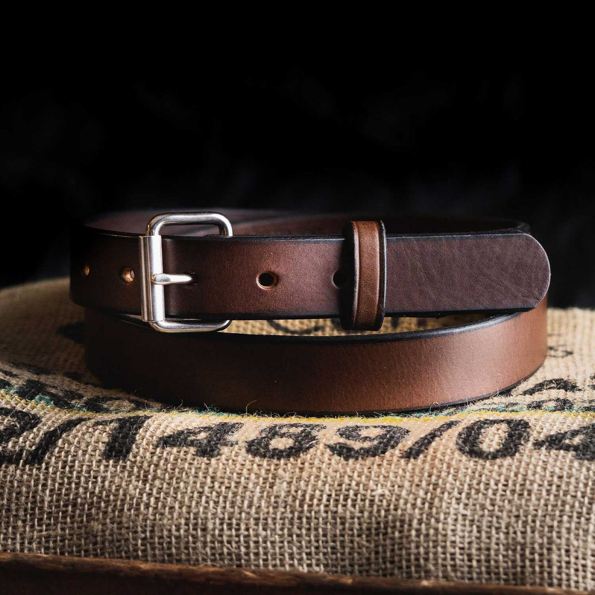 The Deputy Belt - 1.25&quot; from Hanks Belts, featuring a narrow brown leather design with a silver buckle, is coiled and displayed on a textured burlap surface. The dark background accentuates the rich color and durable texture of the belt.