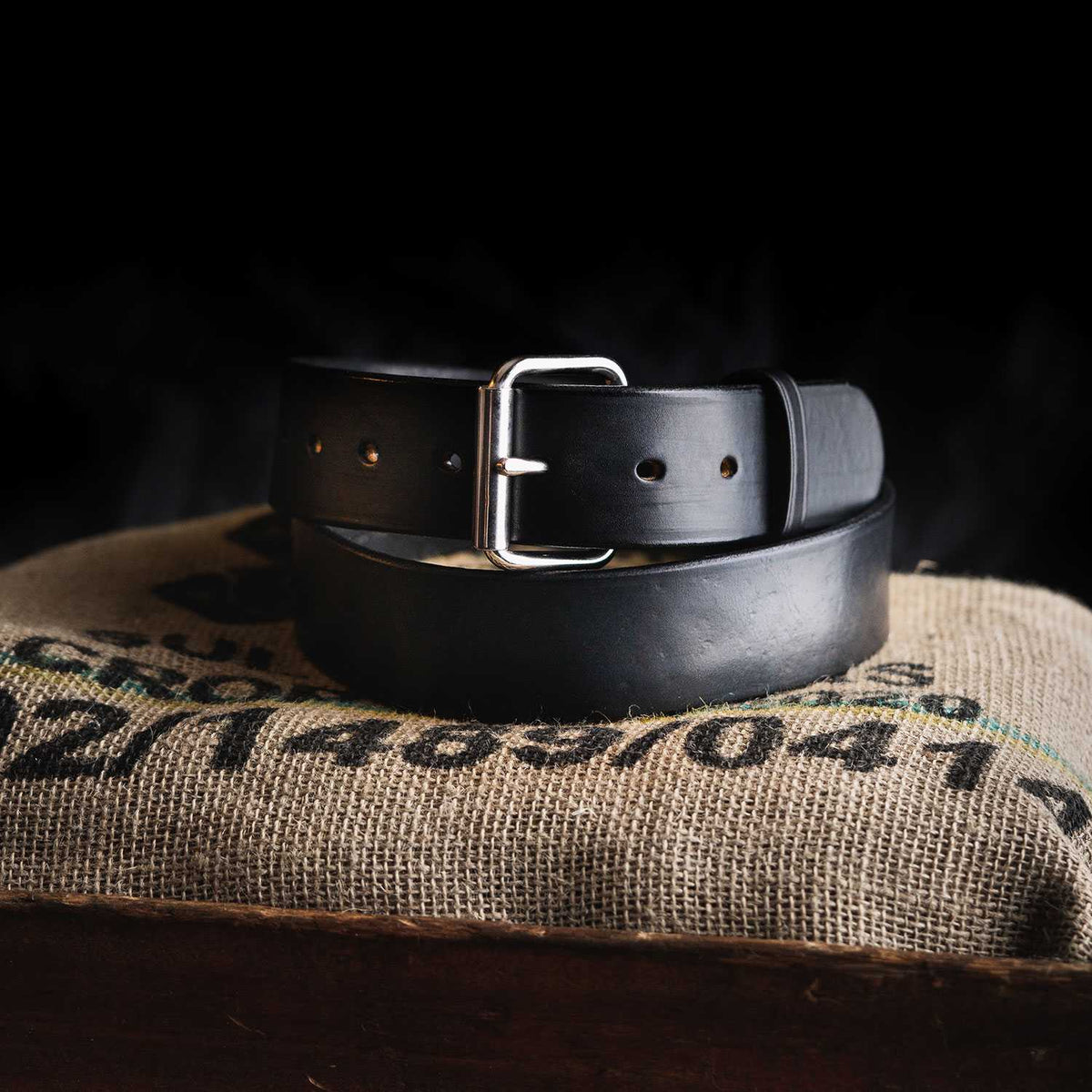 A black leather Enforcer 1.75&quot; gun belt from Hanks Belts, featuring a silver buckle, sits coiled on a piece of textured burlap fabric. The dark background accentuates the sharp contrast of the belt and fabric. The burlap has some indistinct text and numbers printed on it.