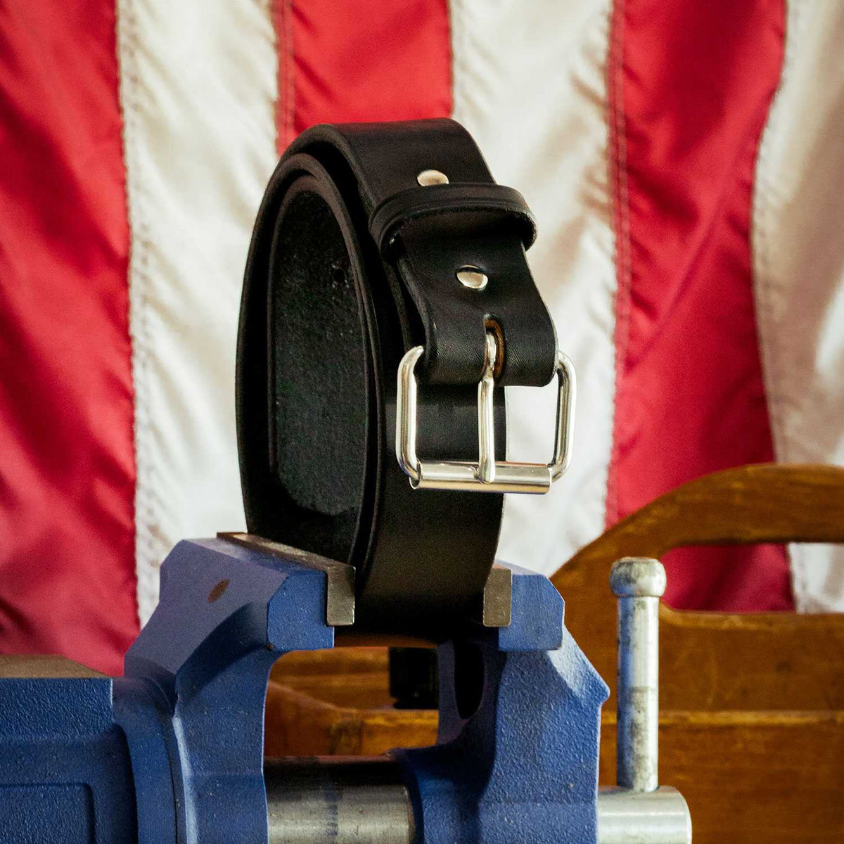 The Enforcer - 1.75&quot; from Hanks Belts, a black leather belt with a shiny silver buckle reminiscent of a LEO&#39;s CCW gun belt, is neatly coiled and held upright in a blue clamp. The background showcases a blurred American flag and part of a wooden object.