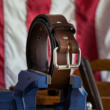 Displayed on a blue vise is "The Enforcer - 1.75"" brown leather belt by Hanks Belts, featuring a silver buckle and resembling an LEO's CCW gun belt. The background highlights a blurred portion of the American flag.