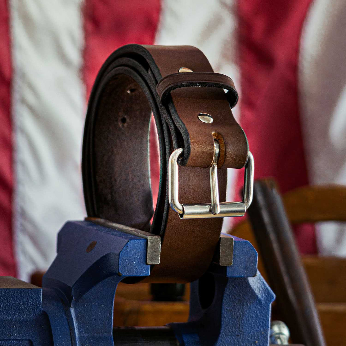Displayed on a blue vise is &quot;The Enforcer - 1.75&quot;&quot; brown leather belt by Hanks Belts, featuring a silver buckle and resembling an LEO&#39;s CCW gun belt. The background highlights a blurred portion of the American flag.