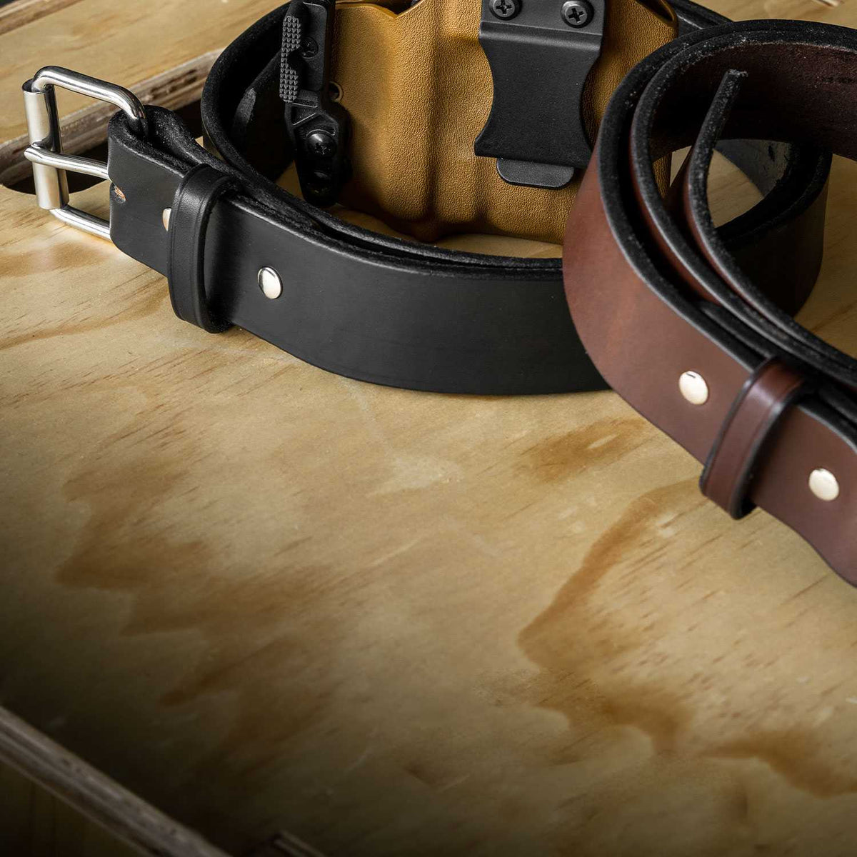 Two leather belts lie on a wooden surface. One belt, named &quot;The Enforcer - 1.75&quot;&quot; by Hanks Belts, is black with a silver buckle, while the other is brown with metal rivets, resembling an LEO&#39;s CCW gun belt. A tan-colored holster with a black clip is partially visible in the background.