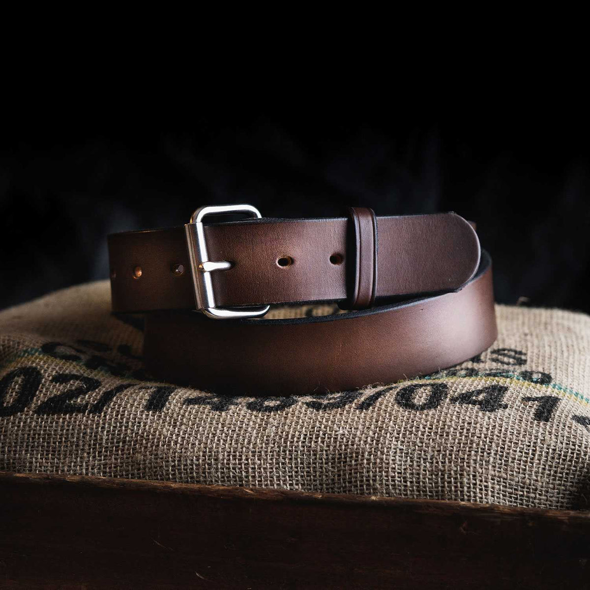 A dark brown Hanks Belts Enforcer 1.75&quot; CCW Gun belt with a silver buckle is coiled neatly atop a piece of burlap fabric. The belt, featuring multiple holes for adjusting the fit, is displayed against a black background. The burlap beneath has faint writing in black and green ink.