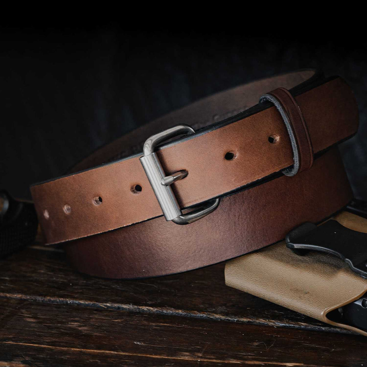 A close-up of &quot;The Gunner - 1.5&quot;&quot; by Hanks Belts, showcasing its silver buckle, lying on a dark wooden surface. Crafted from full-grain leather, this USA Made belt features multiple holes for adjusting the size and a small loop to secure the end. The dark, slightly blurred background beautifully highlights the belt.
