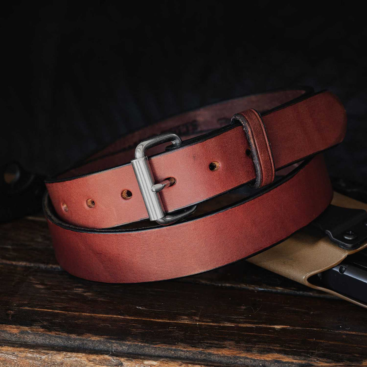 A brown full-grain leather belt from Hanks Belts&#39; &quot;The Gunner - 1.5&quot;&quot; collection is coiled neatly on a wooden surface against a dark background. It features multiple adjustment holes and showcases a natural, polished leather finish. Proudly USA Made, this belt epitomizes quality and craftsmanship with its silver buckle and meticulous design.