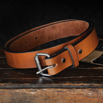 A tan Hanks Belts "The Gunner - 1.5"" belt, crafted from full-grain leather, is laying coiled on a dark wooden surface against a black background. This USA-made belt with a silver buckle has several holes for adjusting the size and features visible stitching along the edges.
