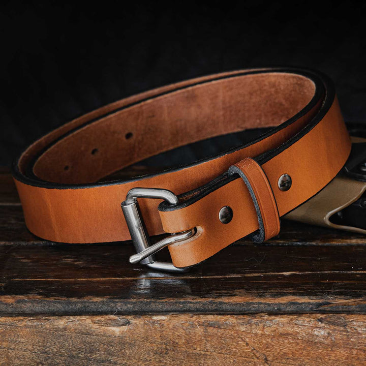 A tan Hanks Belts "The Gunner - 1.5"" belt, crafted from full-grain leather, is laying coiled on a dark wooden surface against a black background. This USA-made belt with a silver buckle has several holes for adjusting the size and features visible stitching along the edges.