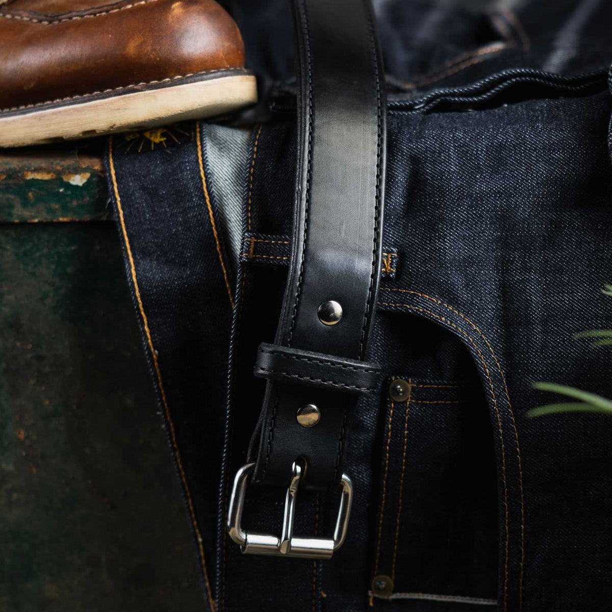 A close-up of a pair of dark blue denim jeans with contrast stitching, laid flat with a black leather Hanks Belts Steel Core Belt - 1.5&quot; featuring a silver buckle draped across them. The edge of a brown leather boot is visible at the top left corner of the image.