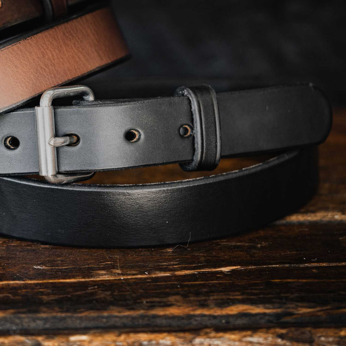 Close-up of two durable leather belts resting on a wooden surface. The top belt is a narrow concealed carry belt, brown with black edges and an antique brass buckle. The bottom belt is Hanks Belts&#39; &quot;The Deputy Belt&quot; in solid black, 1.25&quot; wide with a matte finish and a matching black buckle. The dark background highlights the belts beautifully.