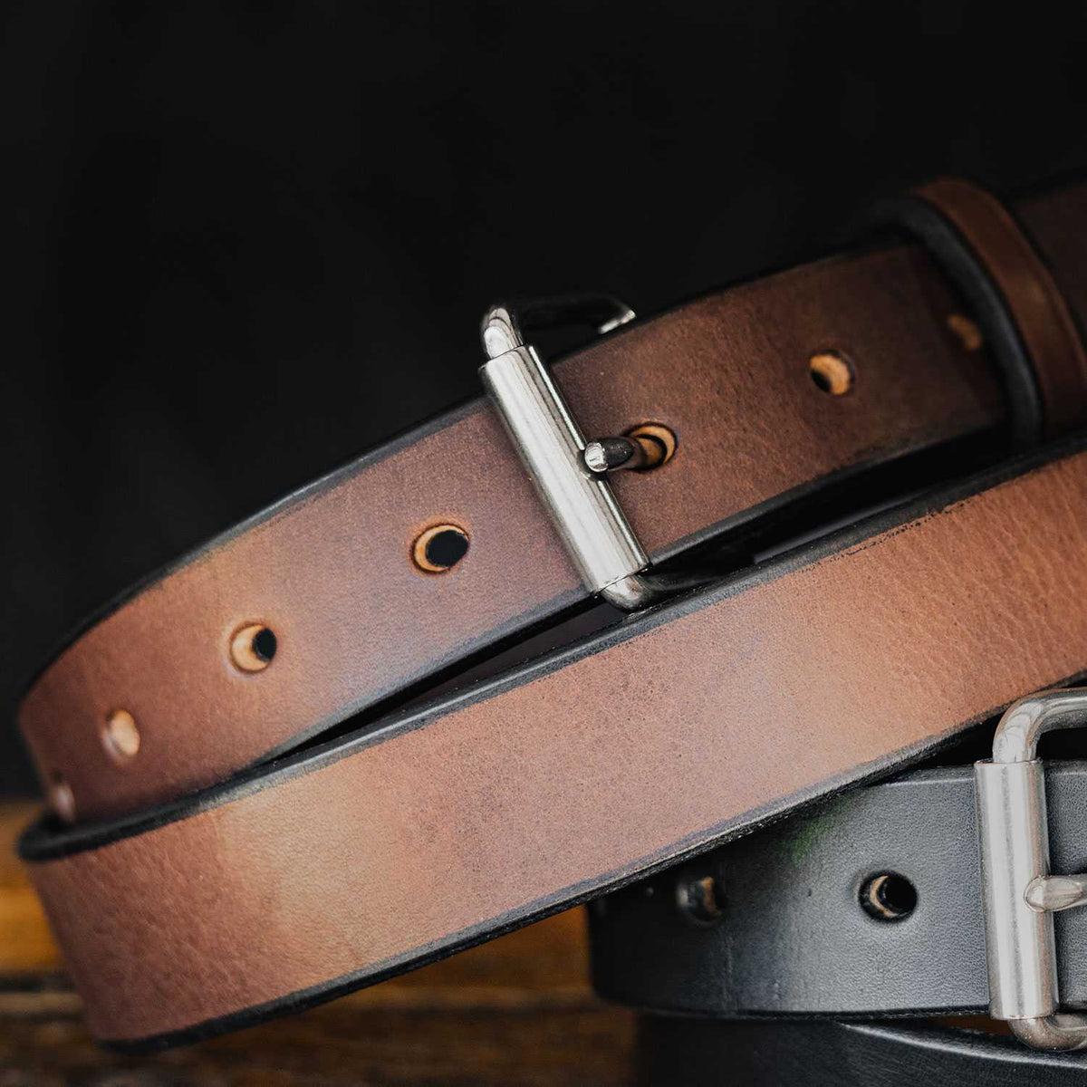 Close-up of two durable leather belts. The top belt, &quot;The Deputy Belt - 1.25&quot;&quot; by Hanks Belts, is brown with a silver buckle and several adjustment holes. The bottom belt is darker, possibly black, with a similar silver buckle and adjustment holes. Both belts are laid on top of each other against a dark background.